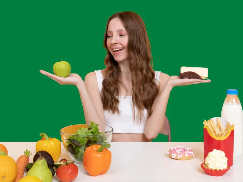 Young white girl holding an apple in her hand and other fruits are placed on that side while also holding a cake in her another hand with junk foods are placed that side